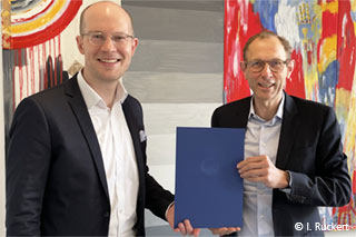 Wiso-Dekan Ulrich W. Thonemann und Juniorporfessor Henrik Franke halten gemeinsam Henrik Frankes Ernennungsurkunde vor einem abstrakten Gemälde.
